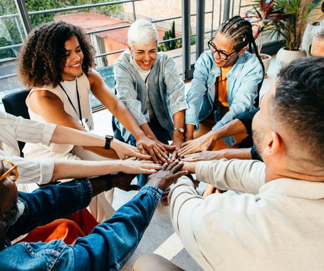 Diverse group of people coming together, placing hands in unity during a team building session in a modern office setting.